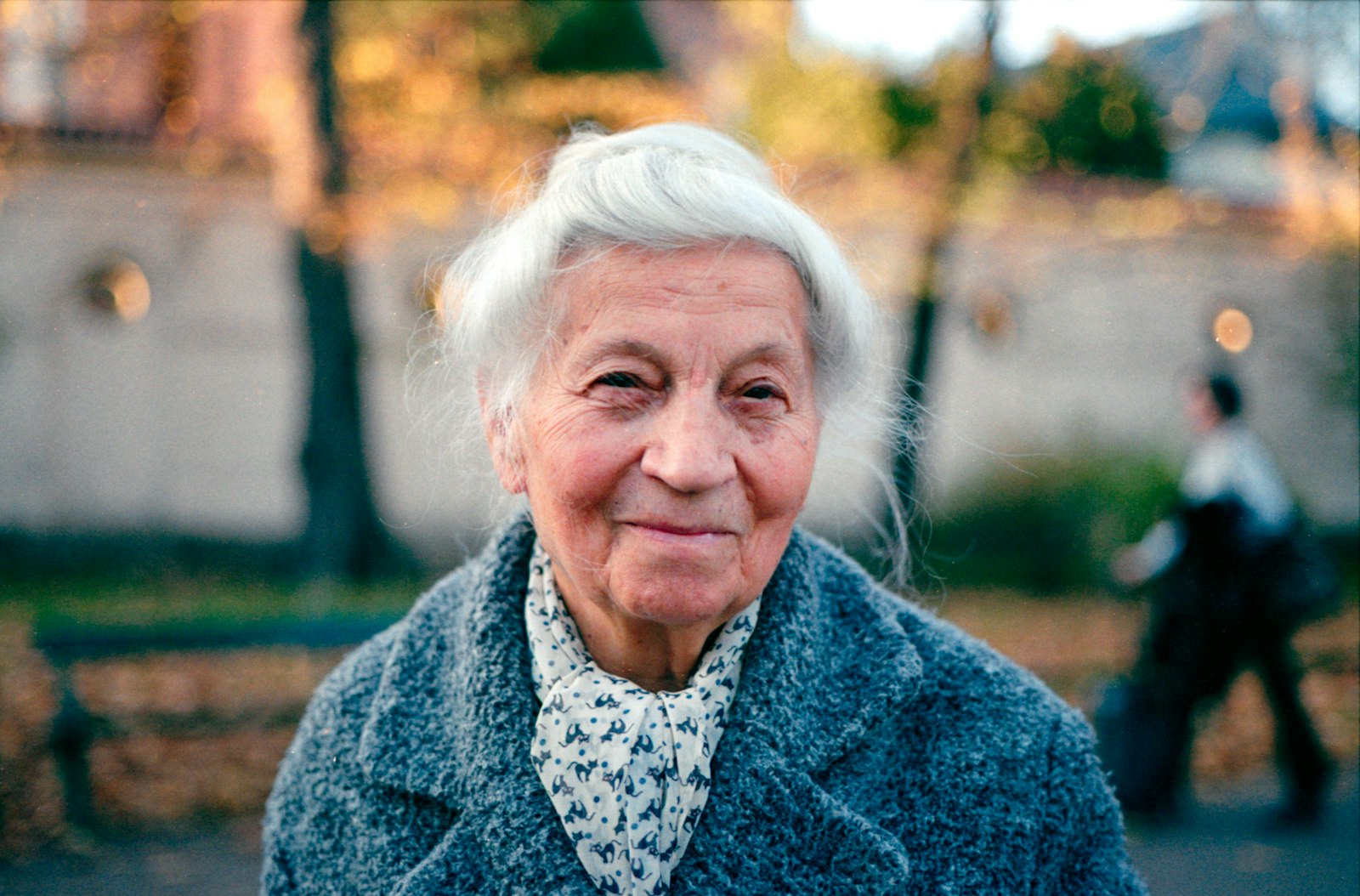 An older woman with white hair in a grey wool coat and patterned scarf, smiling outdoors in warm autumn light.