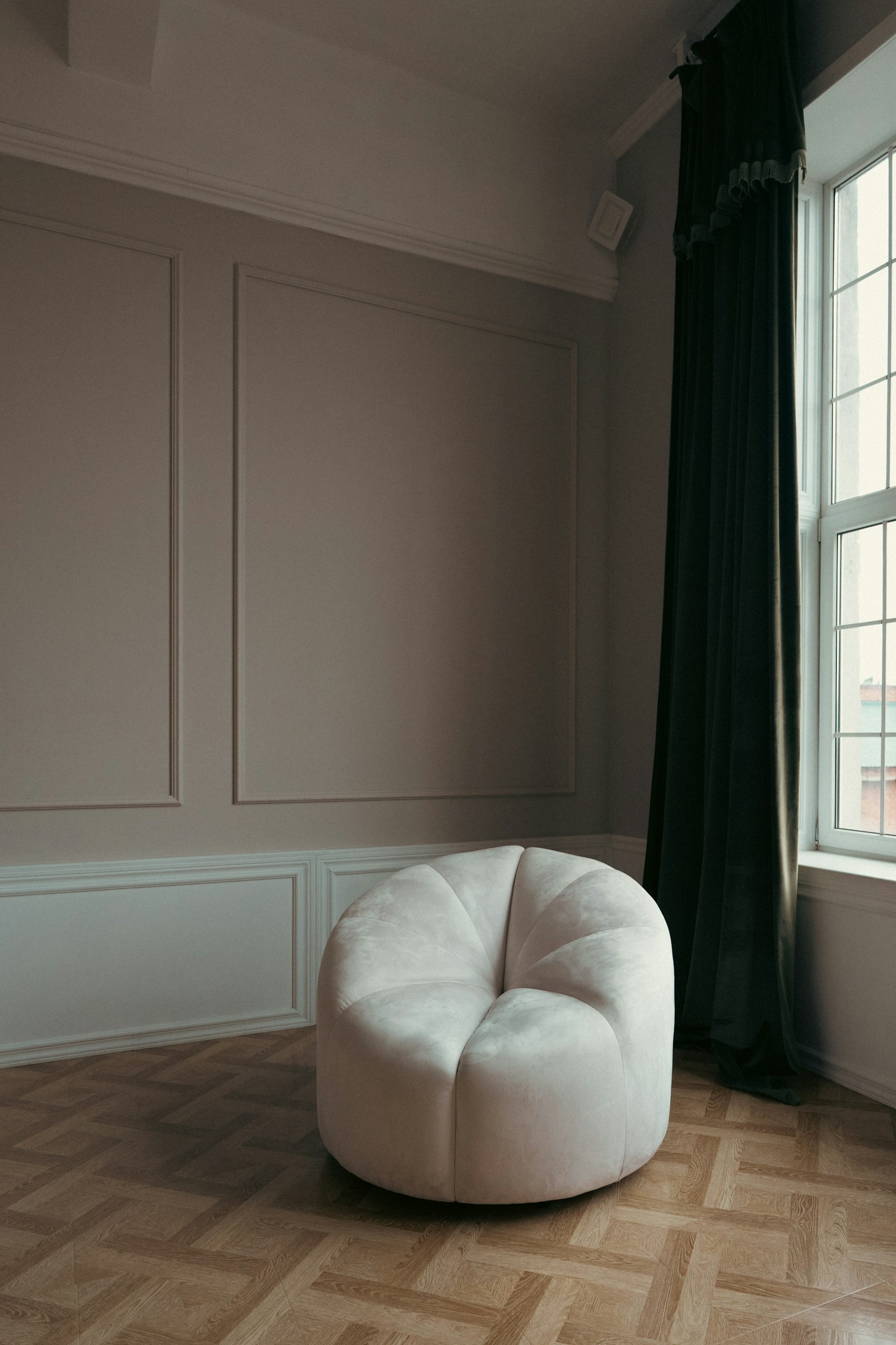 An empty room with pale panelled walls, a parquet floor, a tall curtained window and a single cream armchair.
