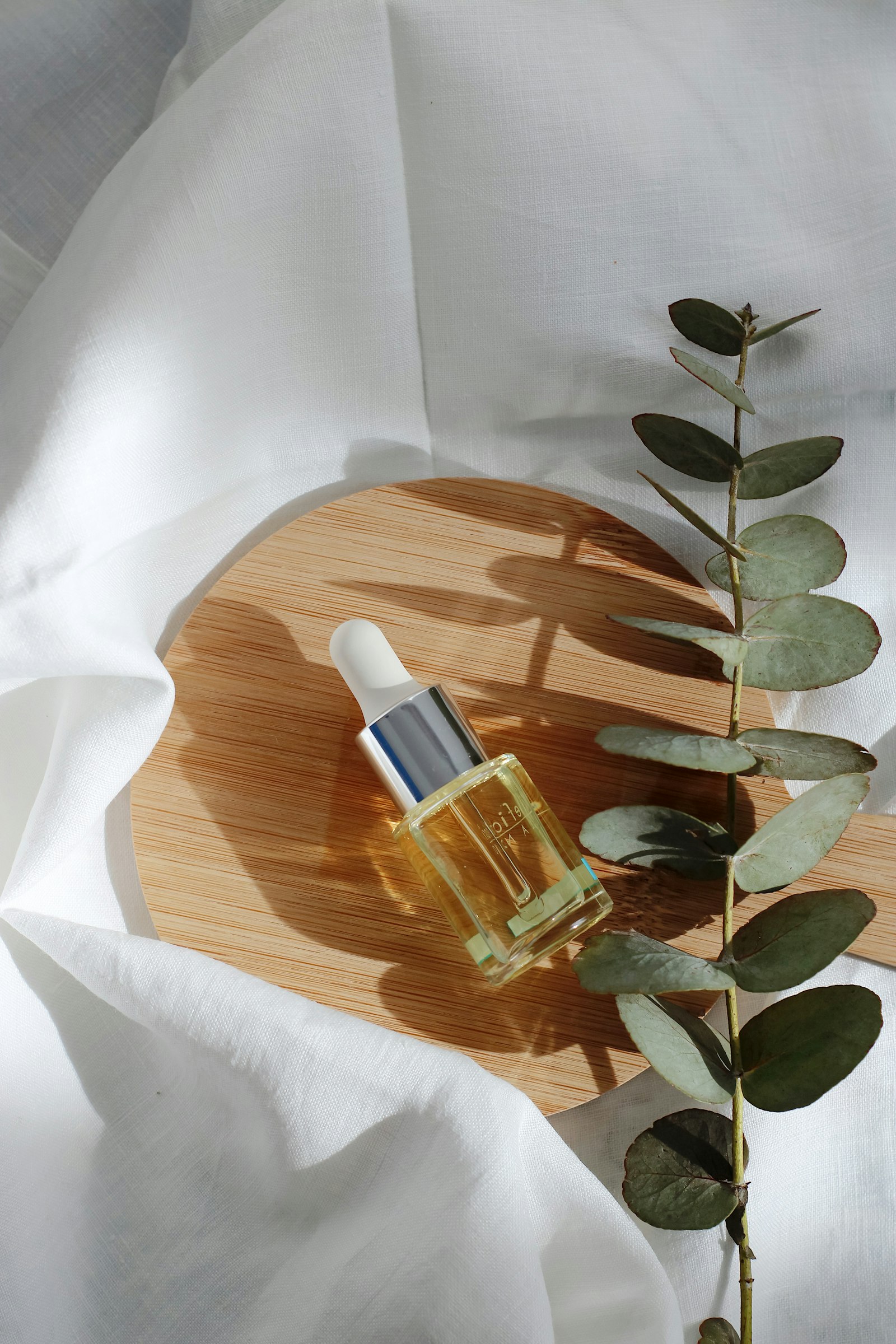 A clear cleansing oil dropper bottle on a bamboo disc beside a eucalyptus sprig