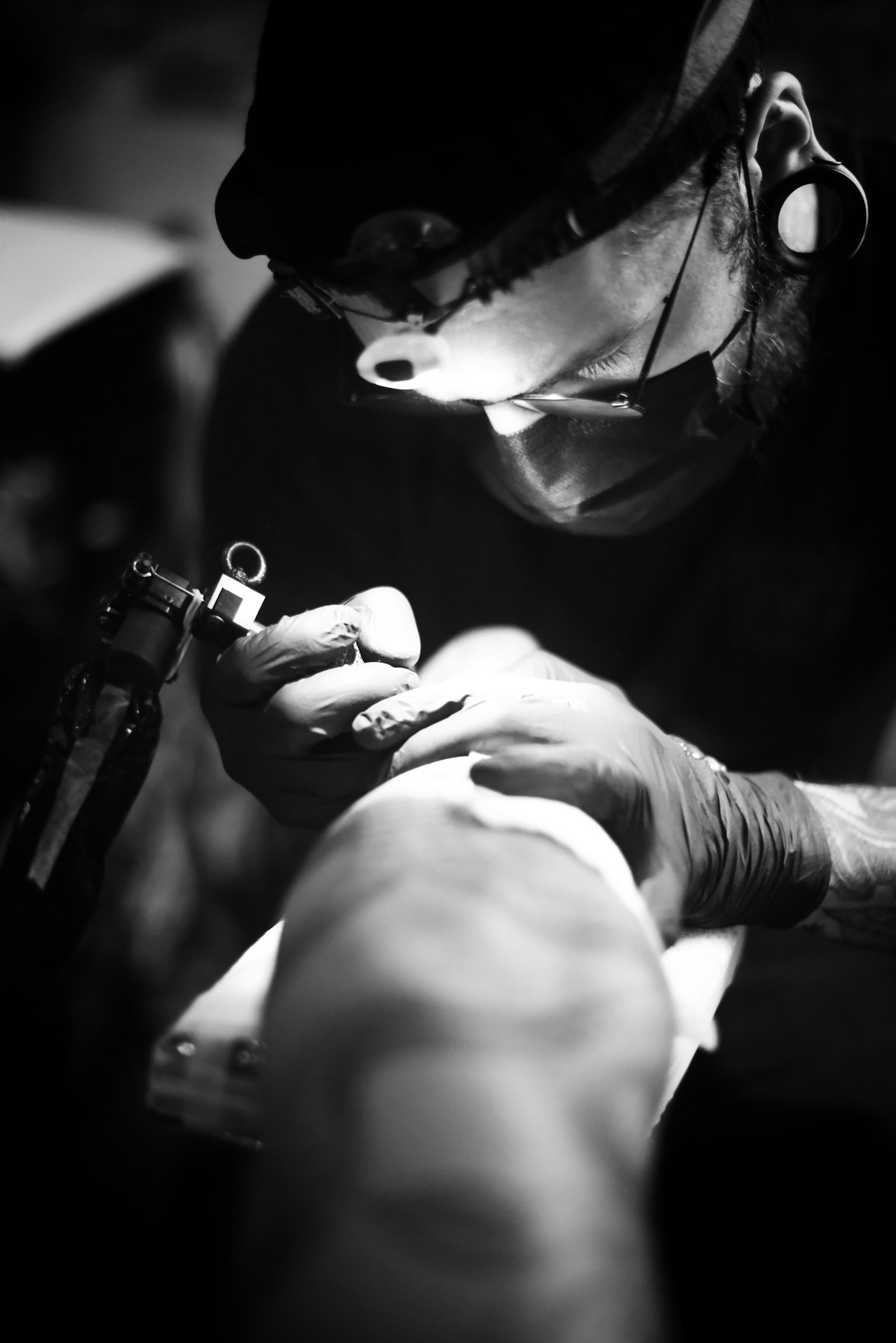 A masked tattoo artist wearing a magnifying loupe leans in close, machine in gloved hand, working under a single hard light