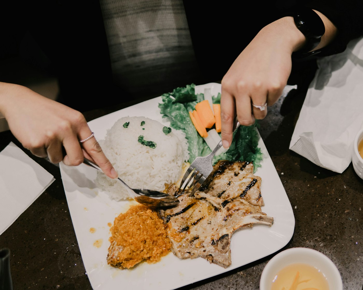 Hai người đang dùng bữa với đĩa cơm sườn nướng, rau xà lách và cà rốt tại bàn ăn văn phòng