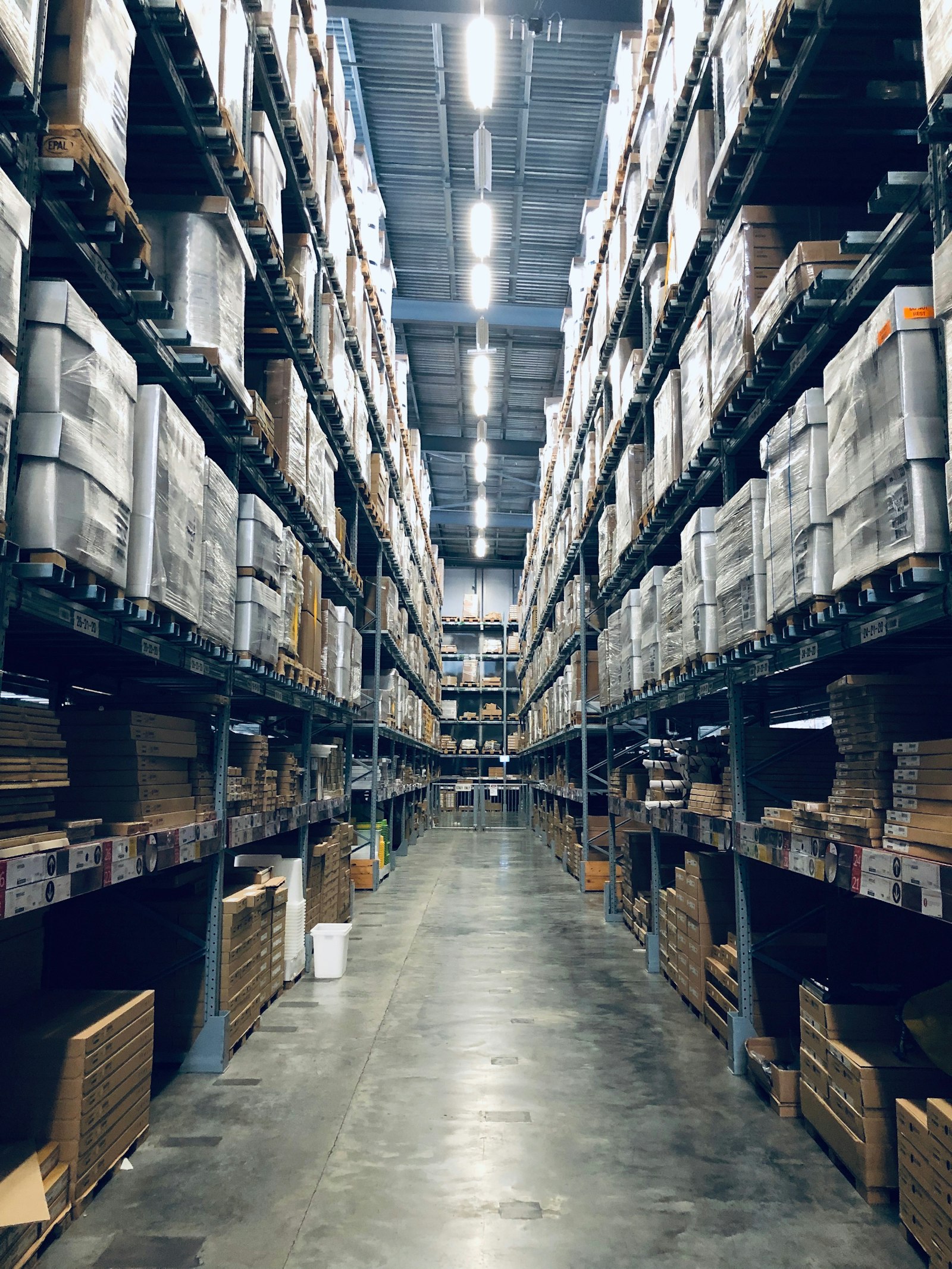 Lối đi giữa hai dãy kệ kho hàng cao chất đầy thùng carton bọc màng, đèn trần sáng dọc lối