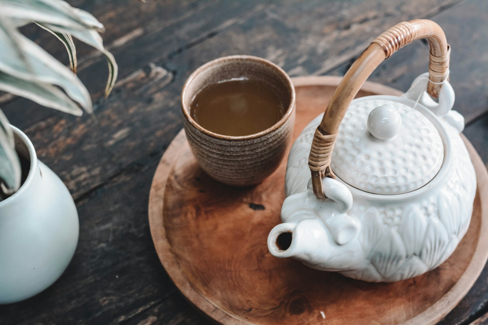 Ấm trà sứ trắng có quai mây và chén trà men nâu đặt trên khay gỗ tròn, mặt bàn gỗ cũ