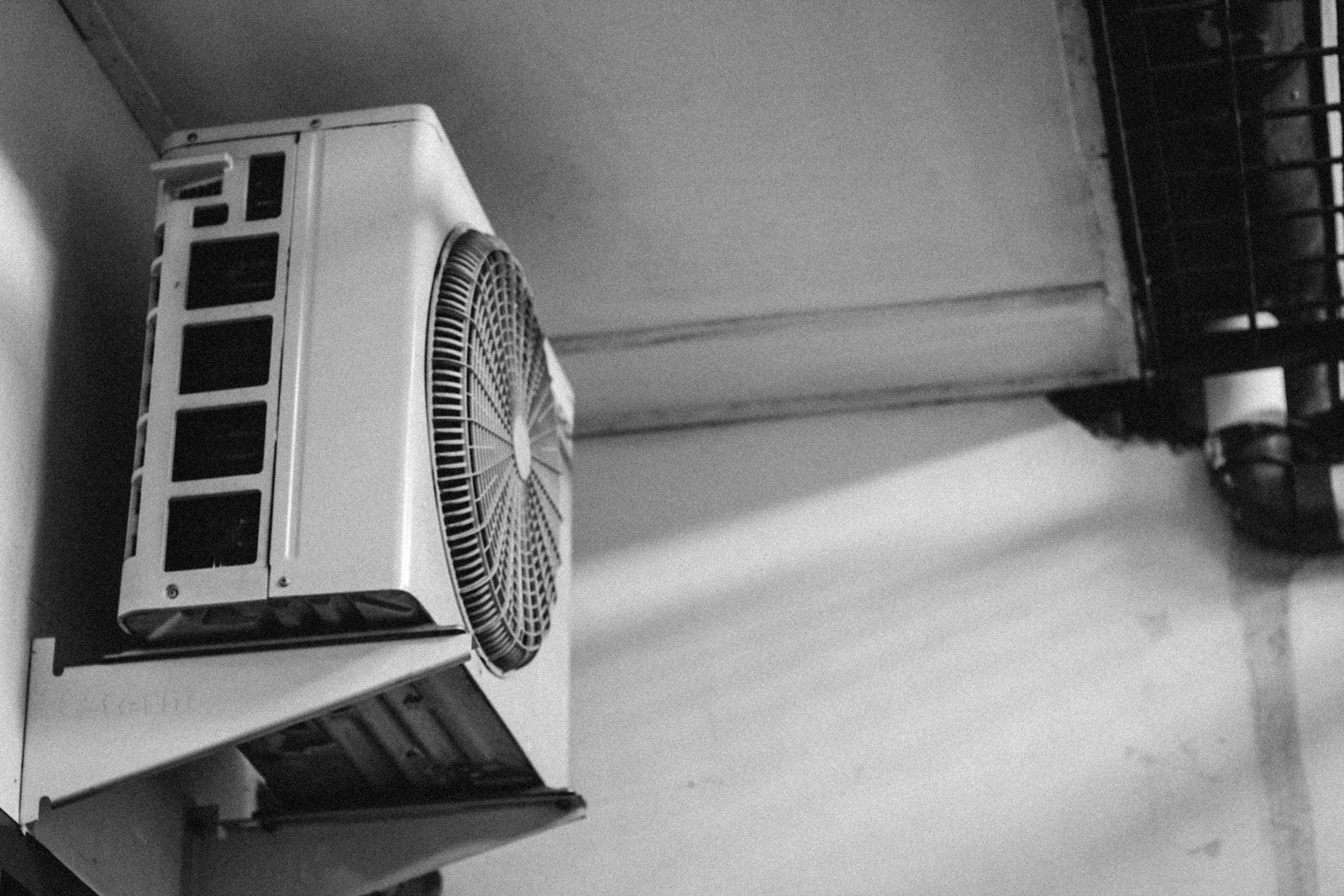 Ảnh trắng đen cụm dàn nóng máy lạnh gắn trên giá đỡ tường, quạt lồng tròn và các khe tản nhiệt.