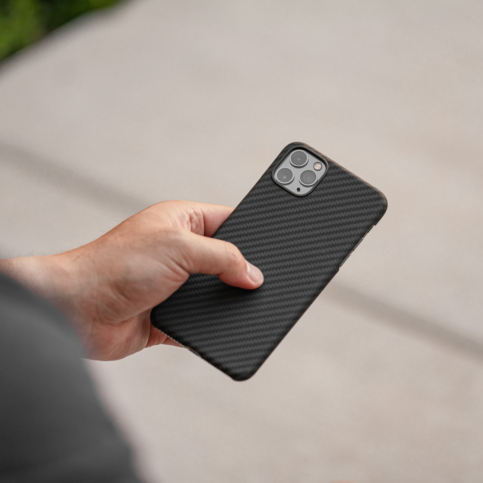 Bàn tay cầm chiếc ốp lưng vân carbon đen nhám, nền vỉa hè sáng mờ phía sau