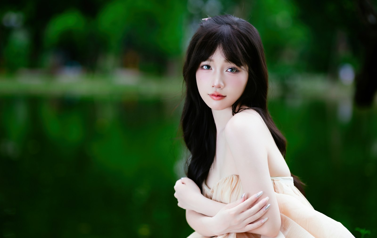 Người phụ nữ tóc dài đen ngồi bên hồ nước, ánh sáng ban ngày dịu, nền cây xanh mờ