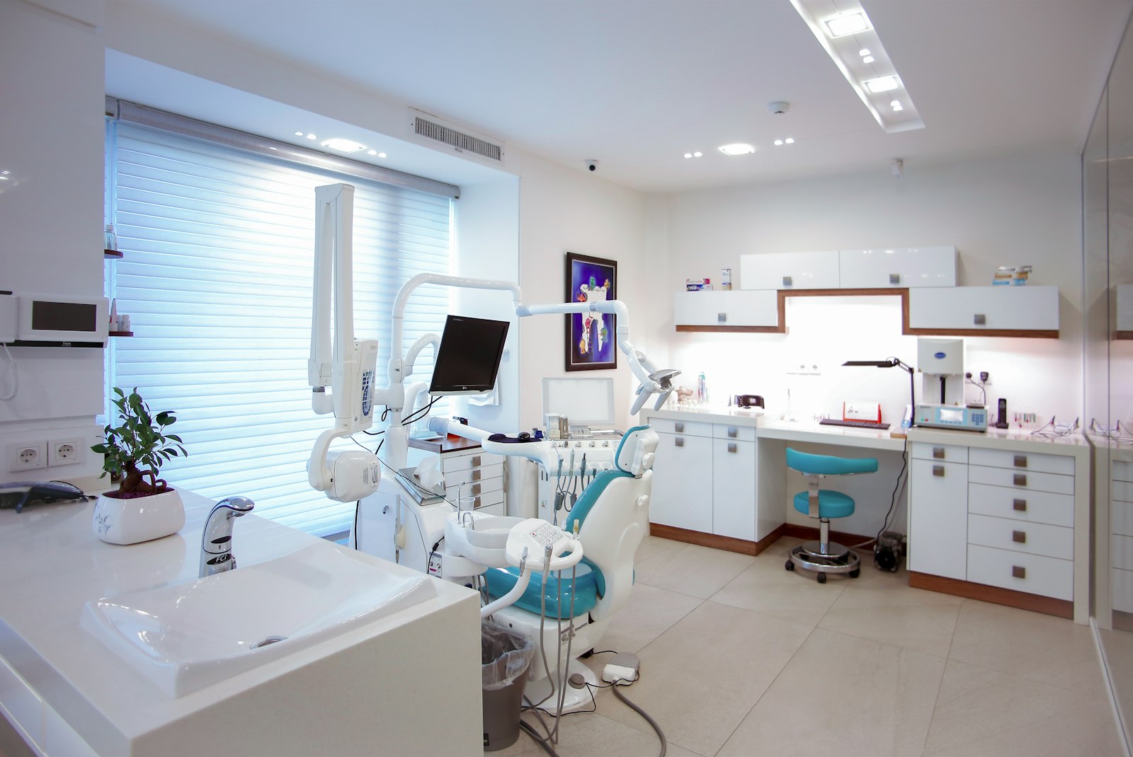 Phòng điều trị nha khoa sáng, ghế nha khoa màu xanh ngọc bên cửa sổ có rèm sáo, tủ trắng và chậu cây nhỏ