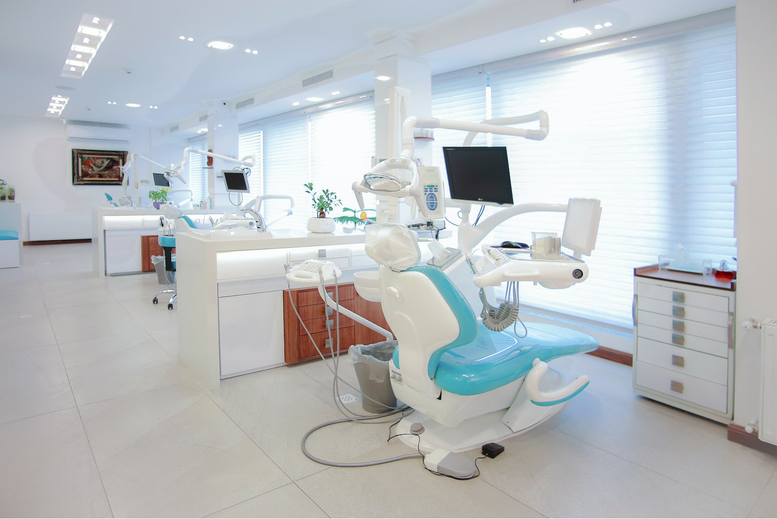 Khu điều trị rộng nhiều ghế nha khoa bọc xanh ngọc xếp thẳng hàng, tủ trắng, cửa sổ có rèm sáo cho ánh sáng vào