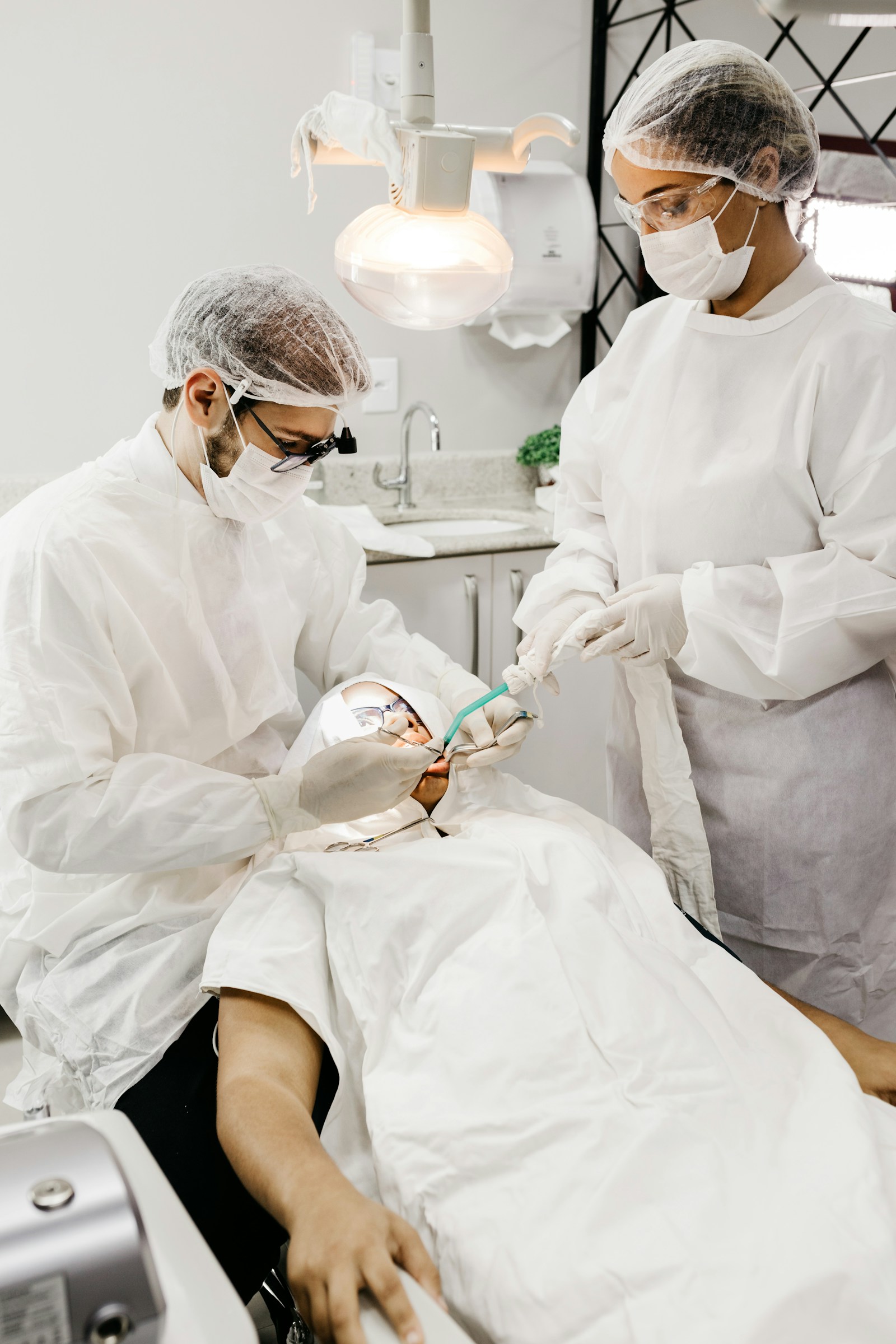 Hai nhân viên y tế mặc đồ bảo hộ trắng kín, đội mũ trùm và đeo khẩu trang, đang cùng làm việc trên một bệnh nhân đã phủ khăn vô trùng