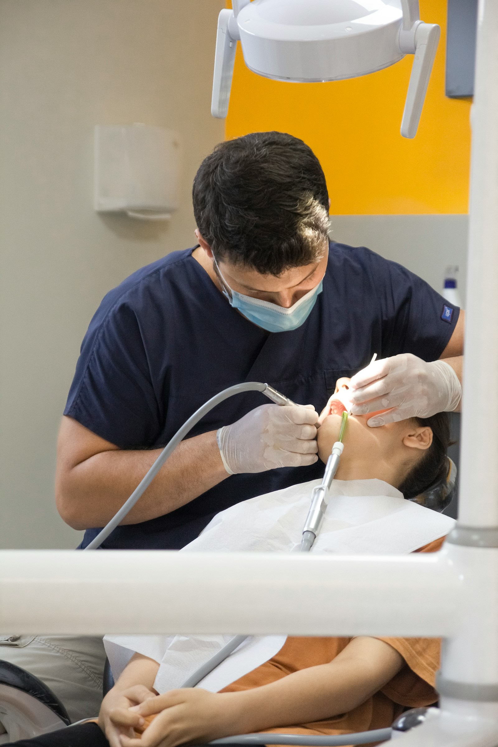 Bác sĩ mặc áo scrub xanh than đang điều trị cho bệnh nhân nằm trên ghế, tay cầm tay khoan và ống hút nước bọt