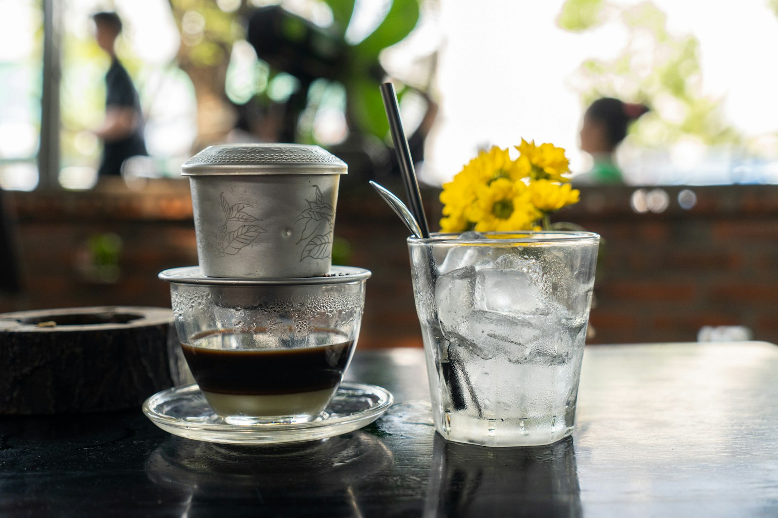 Phin nhôm đặt trên ly cà phê đen pha cùng sữa đặc, bên cạnh là ly đá và bình hoa cúc vàng trên mặt bàn gỗ sẫm