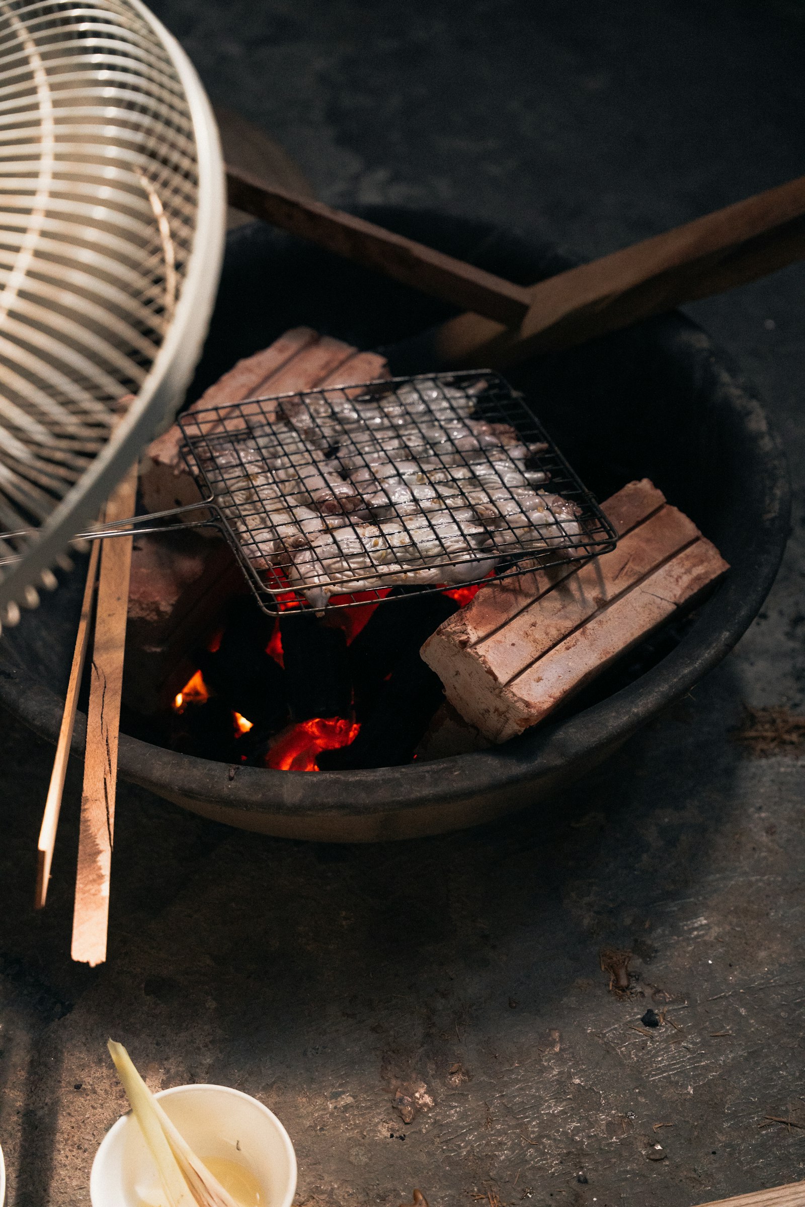 Nhìn từ trên xuống bếp than đất nung, vỉ lưới thép đựng mực nhỏ đang nướng, kẹp tre và gạch chèn quanh lò