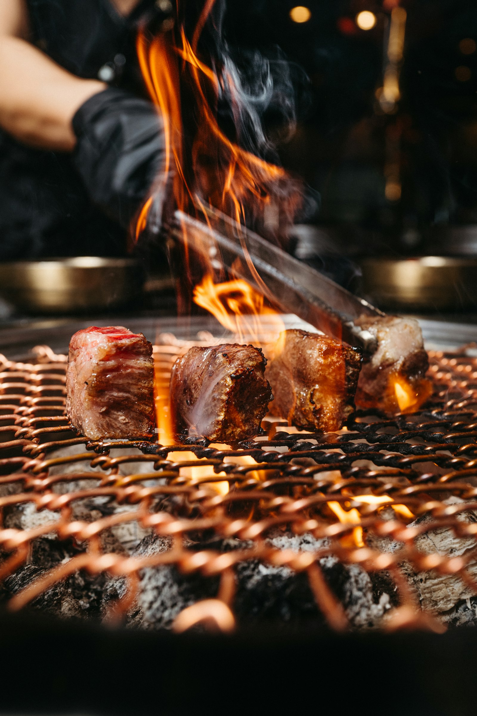 Bốn khối thịt bò dày đang nướng trên vỉ đồng, lửa bùng lên qua kẽ vỉ, kẹp gắp thép phía trên