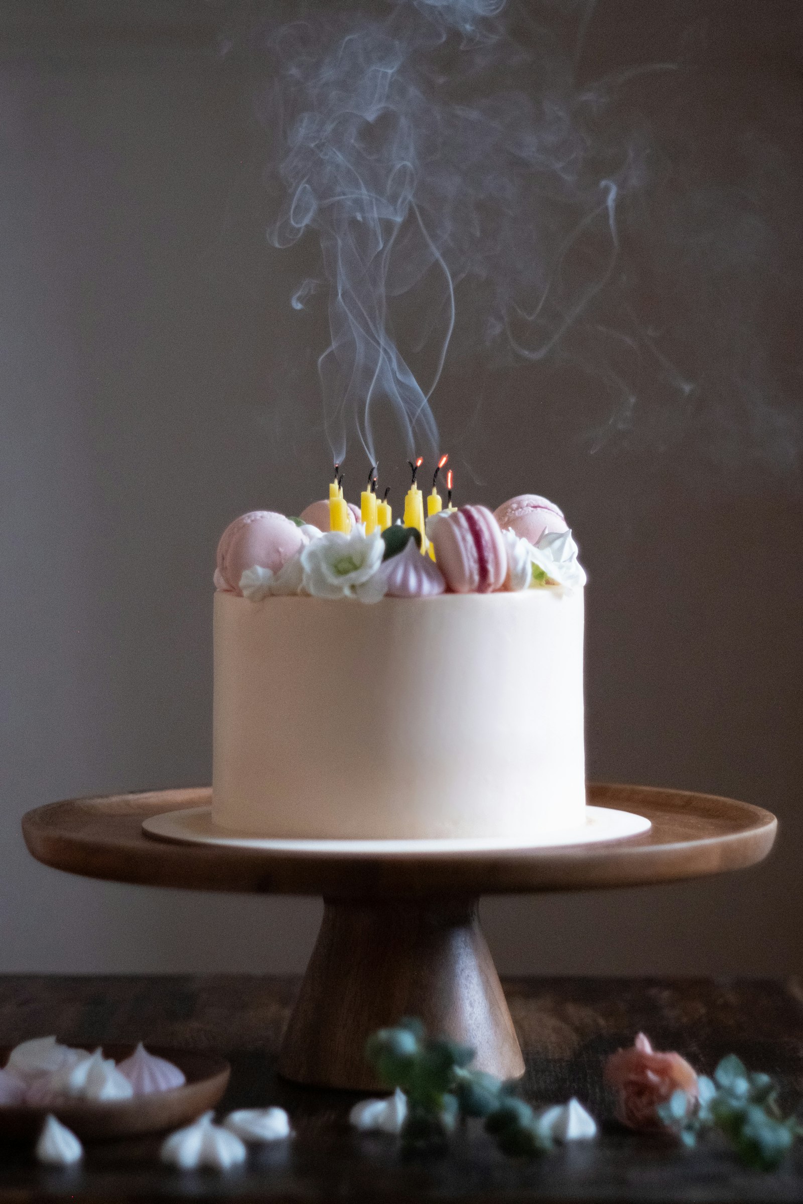 Bánh kem tròn phủ kem trắng ngà, mặt bánh xếp macaron hồng, hoa trắng và bánh trứng đường, cắm nến vàng vừa thổi còn khói.