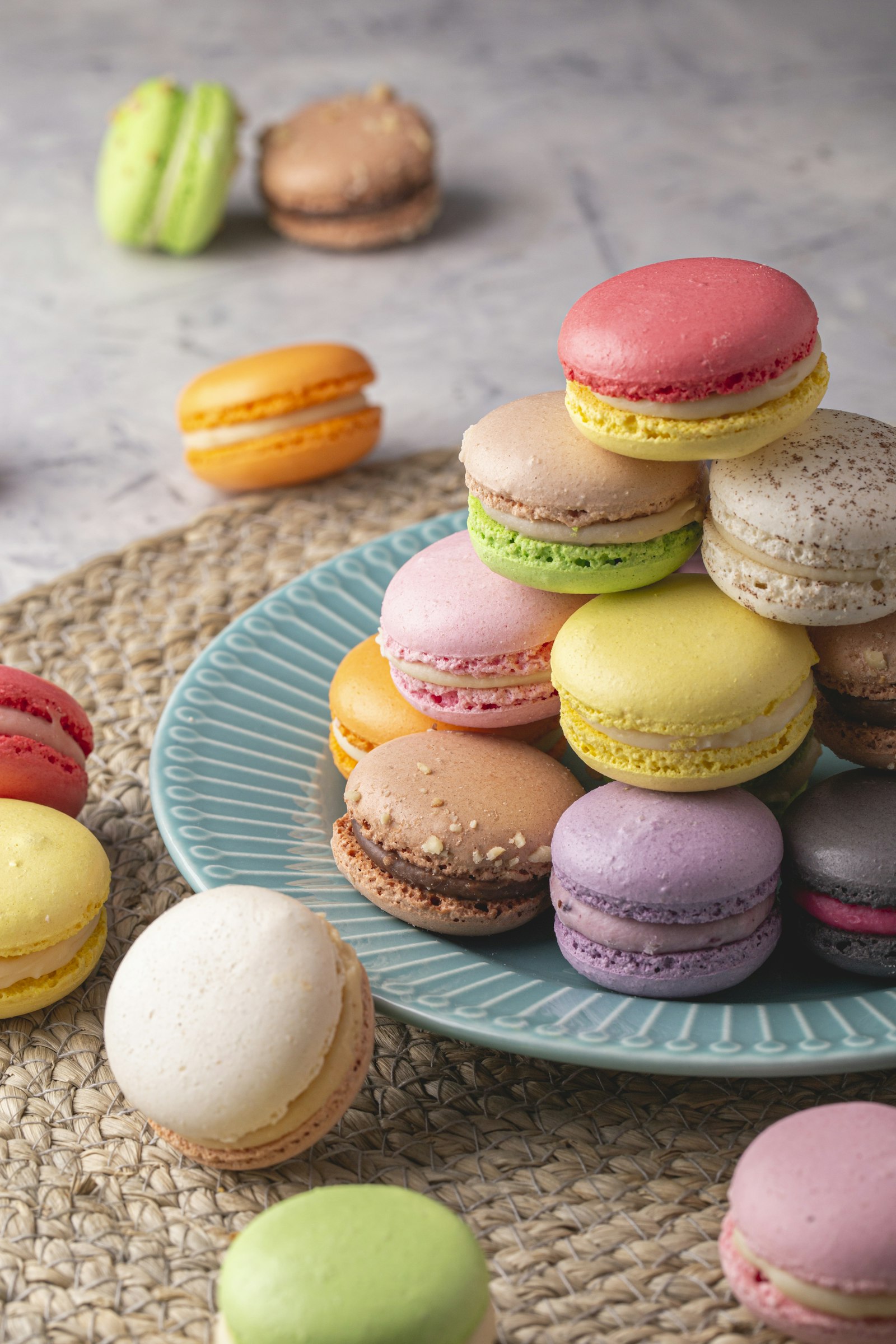 Chồng macaron nhiều màu — hồng, vàng, tím, xanh lá, nâu — xếp trên đĩa sứ xanh ngọc đặt trên tấm lót cói.