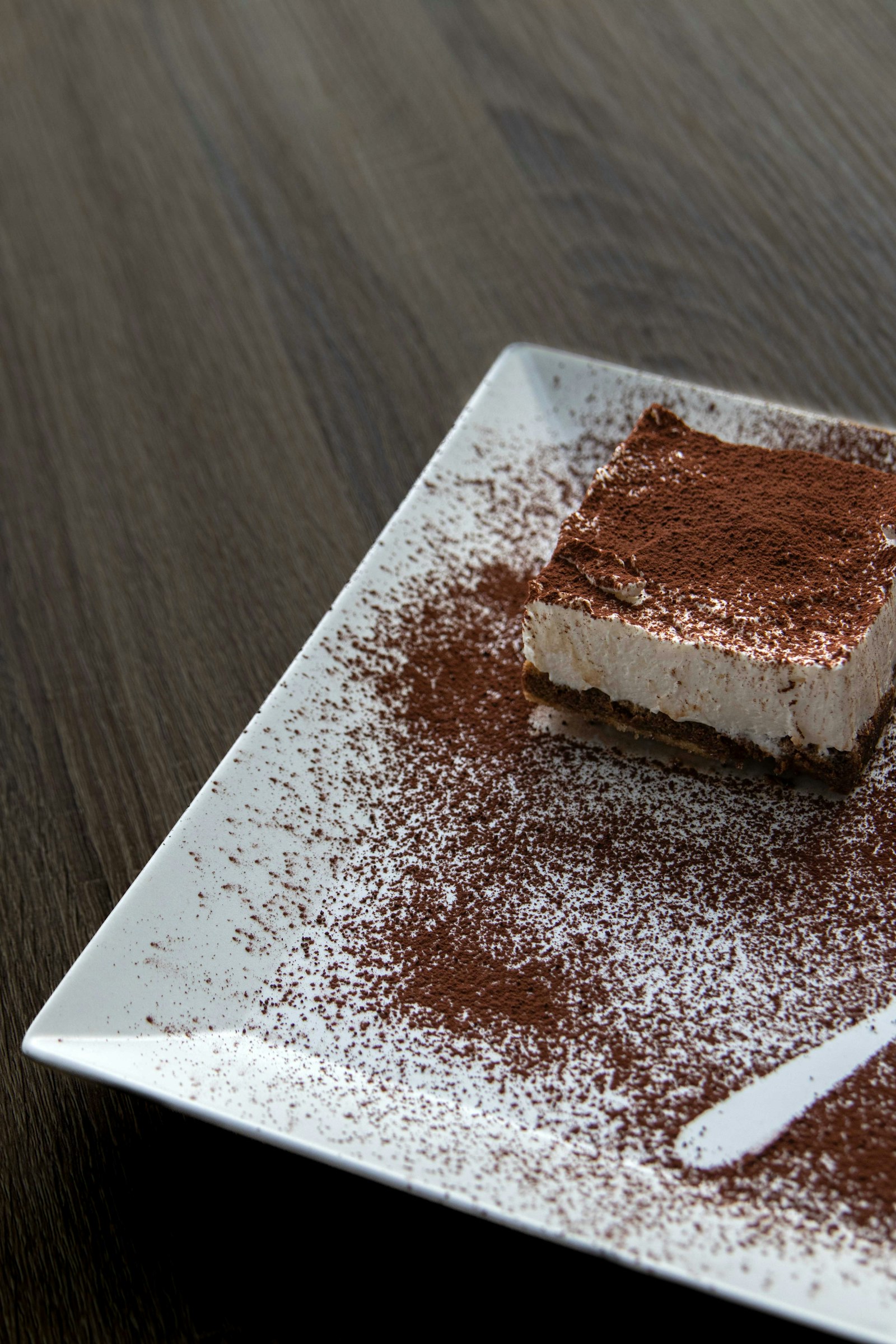 Miếng tiramisu phủ bột cacao trên đĩa chữ nhật trắng.