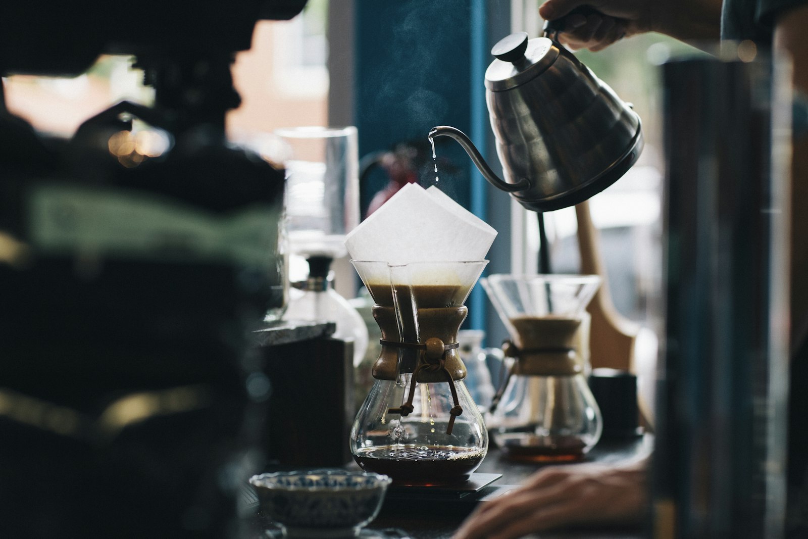 Một bàn tay cầm ấm cổ ngỗng bằng thép rót nước sôi bốc hơi vào phễu lọc giấy đặt trên bình thủy tinh cổ eo, cà phê đã chảy xuống đáy bình, phía sau là cửa sổ sáng.