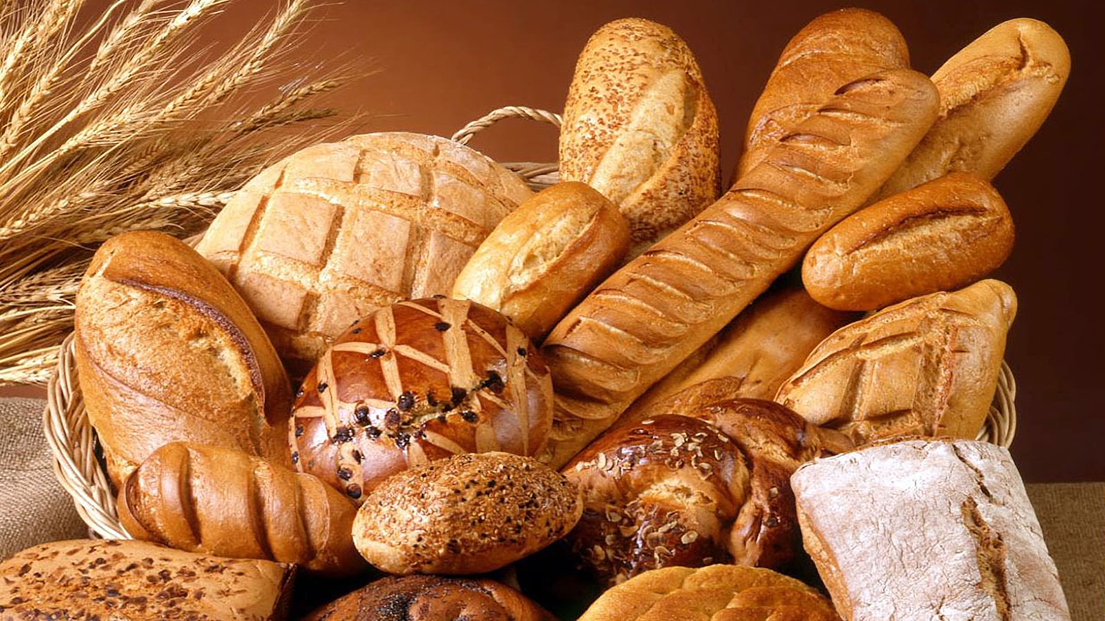 Một rổ mây đầy các loại bánh mì vỏ vàng nâu: bánh mì gậy dài rạch xéo, ổ tròn rắc mè, ổ vuông phủ bột, xếp chồng lên nhau bên cạnh mấy bông lúa mì khô.