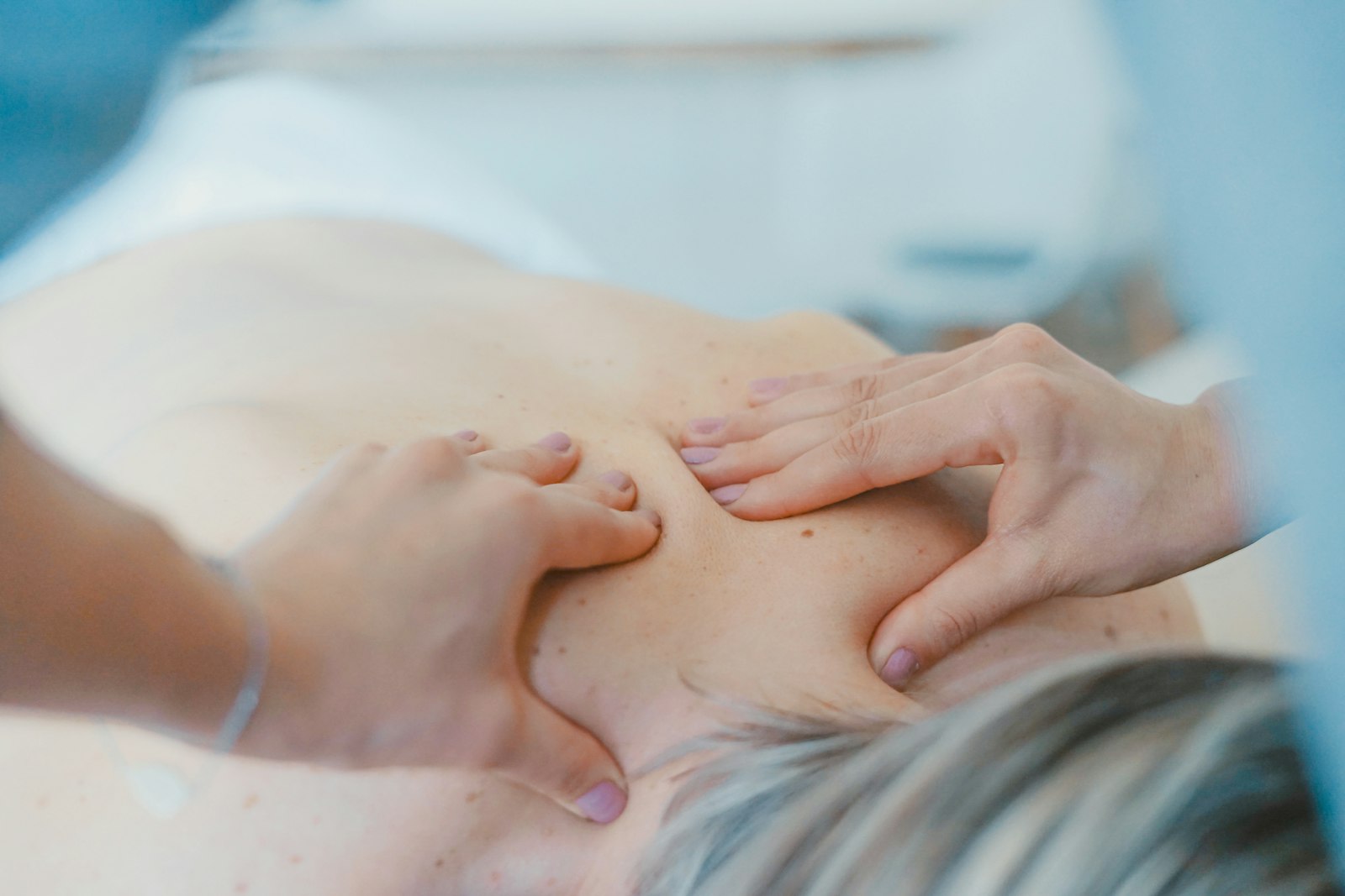 Cận cảnh hai bàn tay kỹ thuật viên đang day ấn vùng vai và bả vai của một người nằm sấp trên giường trải khăn trắng, ánh sáng xanh dịu.