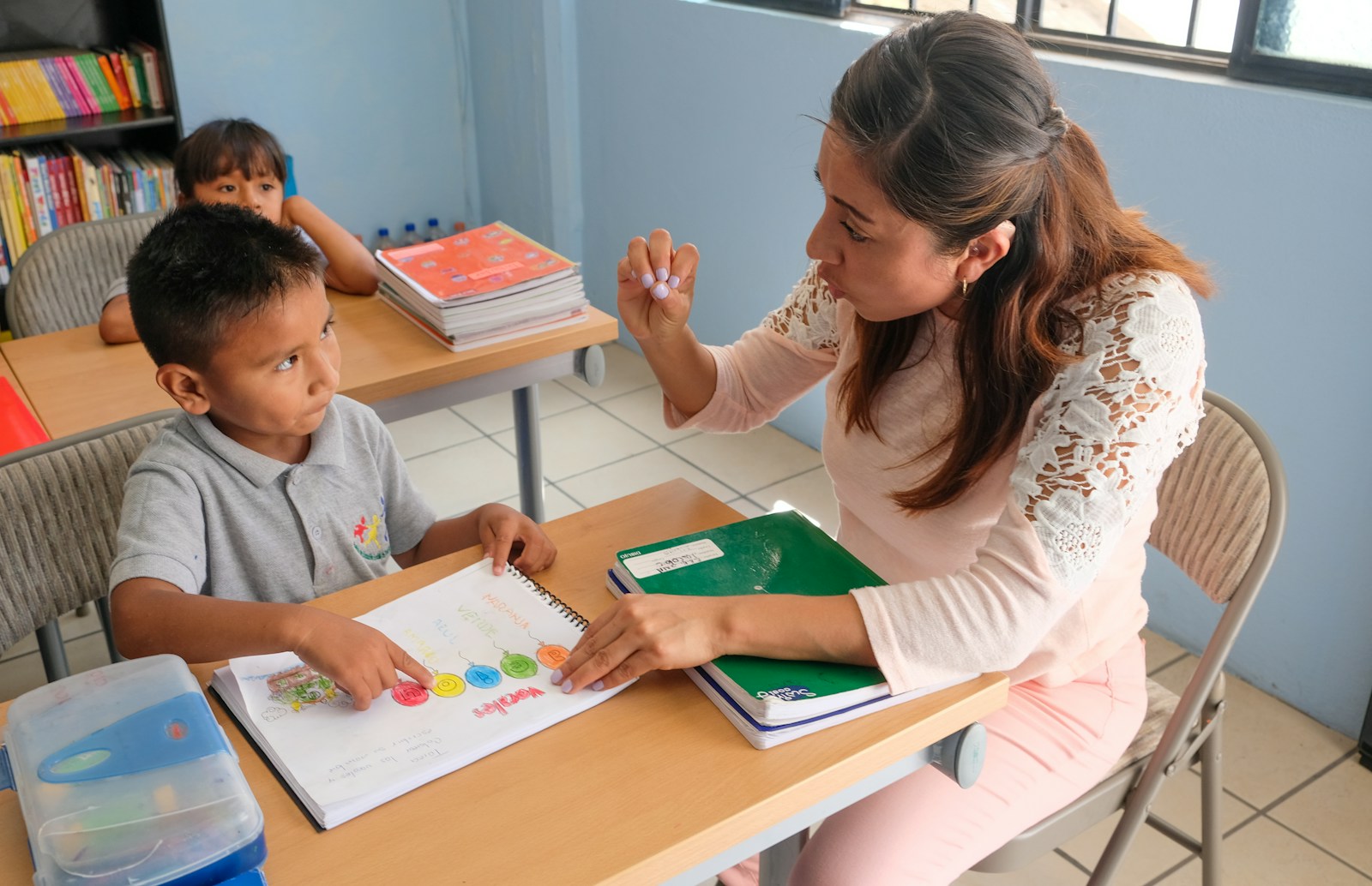 Cô giáo ngồi cạnh bàn hướng dẫn một em nhỏ đang chỉ vào vở bài tập nhiều màu